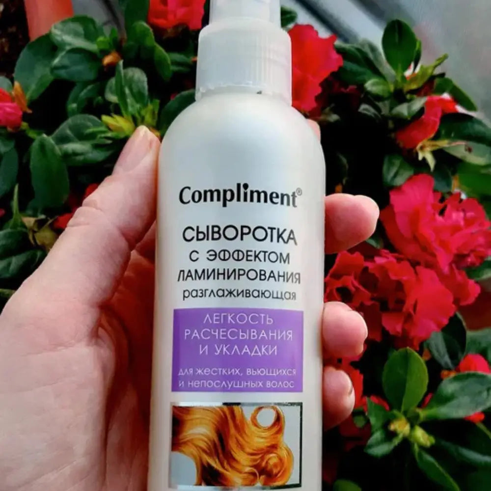 Hand holding a bottle of 'Compliment' hair serum with flowers in the background